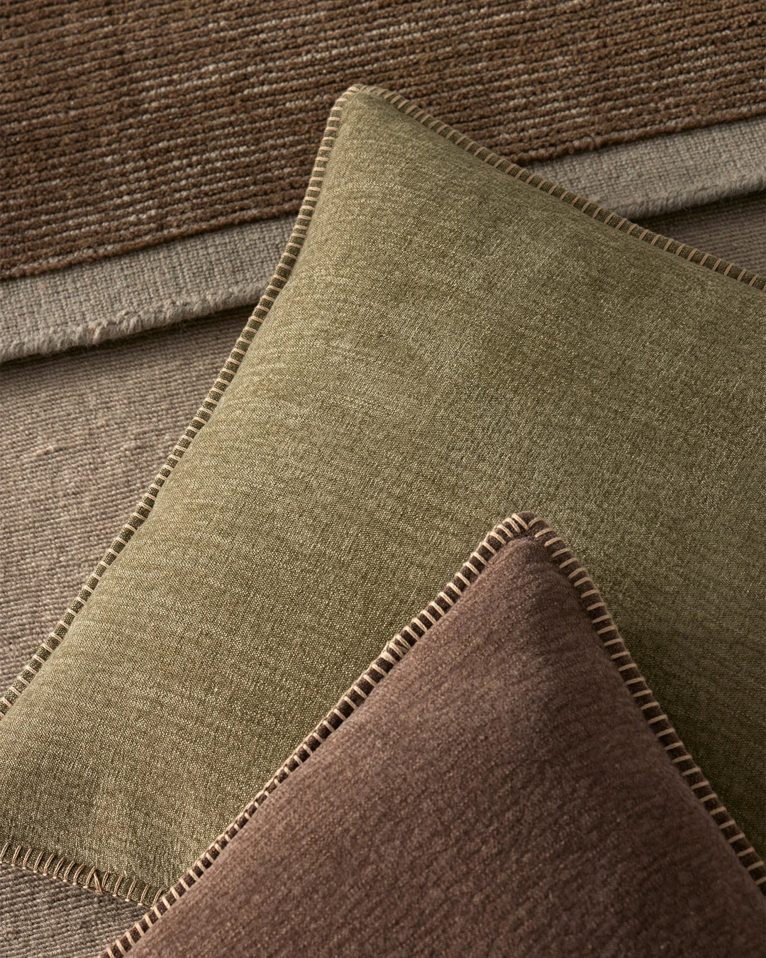 Earthy neutral cushions in sage and chocolate tones layered with textured linens, showcasing blanket-stitch details and natural fibre texture palette