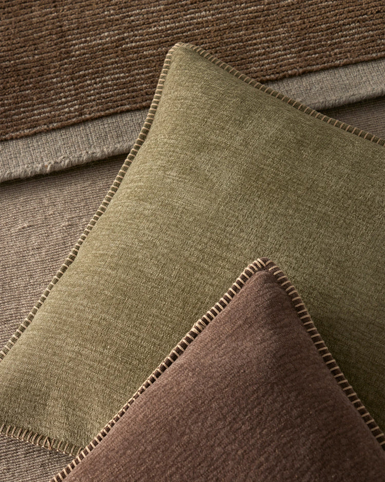 Earthy neutral cushions in sage and chocolate tones layered with textured linens, showcasing blanket-stitch details and natural fibre texture palette