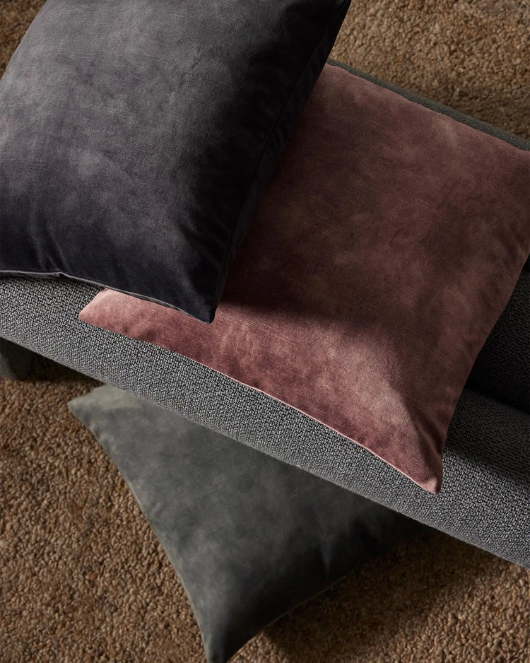 Charcoal and burgundy velvet cushions layered on a grey upholstered sofa with warm taupe textured rug, showcasing moody jewel-tone interior styling