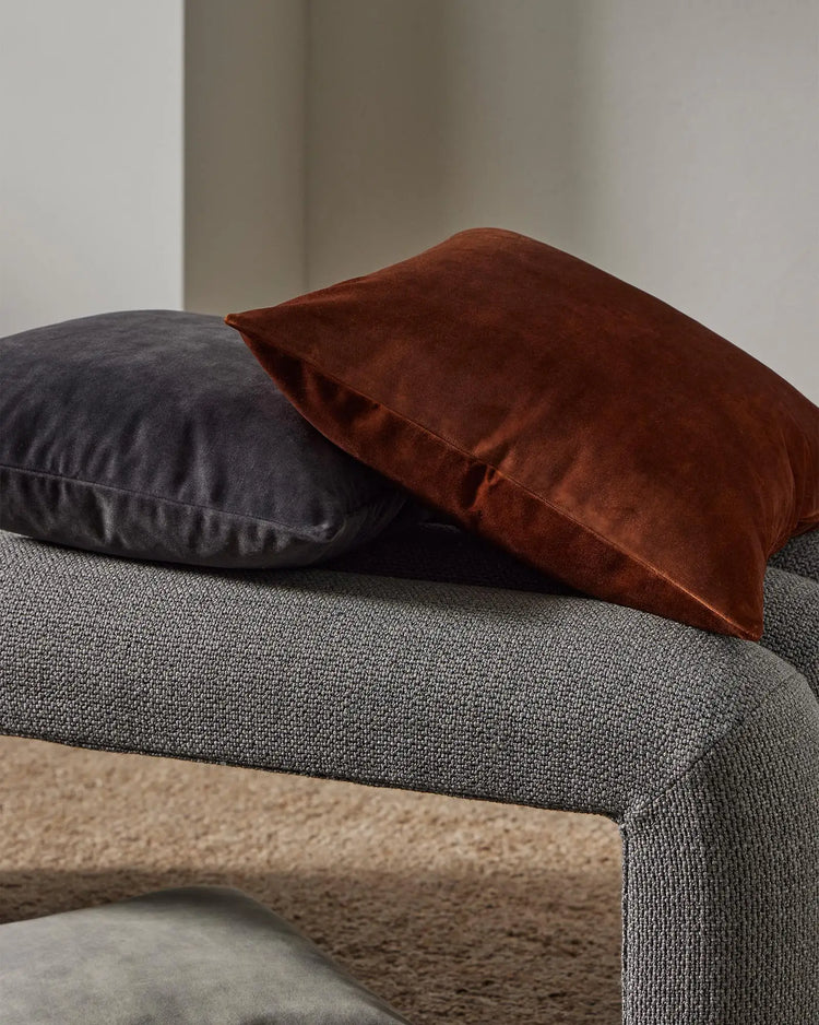 Warm earth-tone cushions in cinnamon and charcoal velvet layered on a grey upholstered sofa, textured throw detail visible against neutral interior backdrop