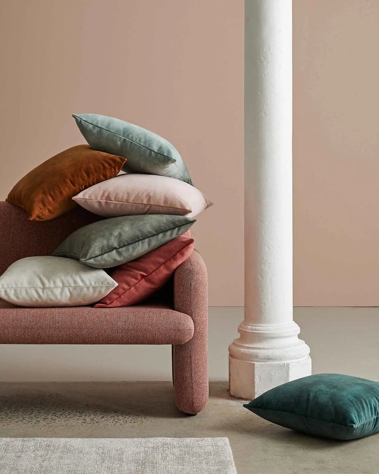 Stacked velvet cushions in ochre, blush, and cinnamon tones layered on a terracotta upholstered chair, styled against soft pink walls and architectural white column for warm, earthy interior design inspiration.