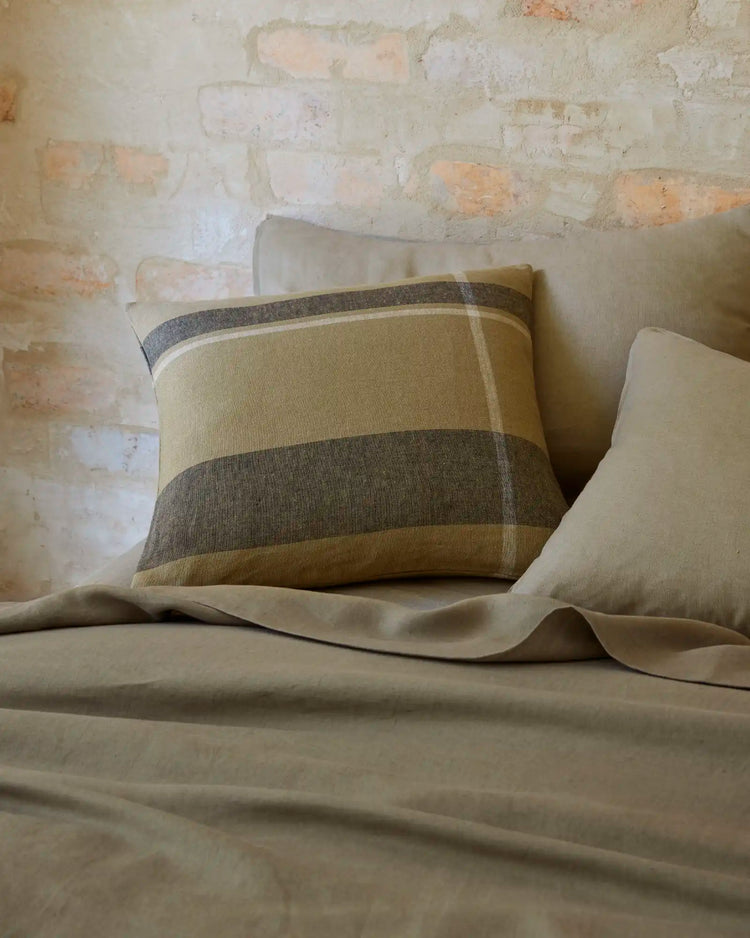 Earthy striped cushion in sage and charcoal on natural linen Ravello bedding, styled against whitewashed brick with soft fabric draping