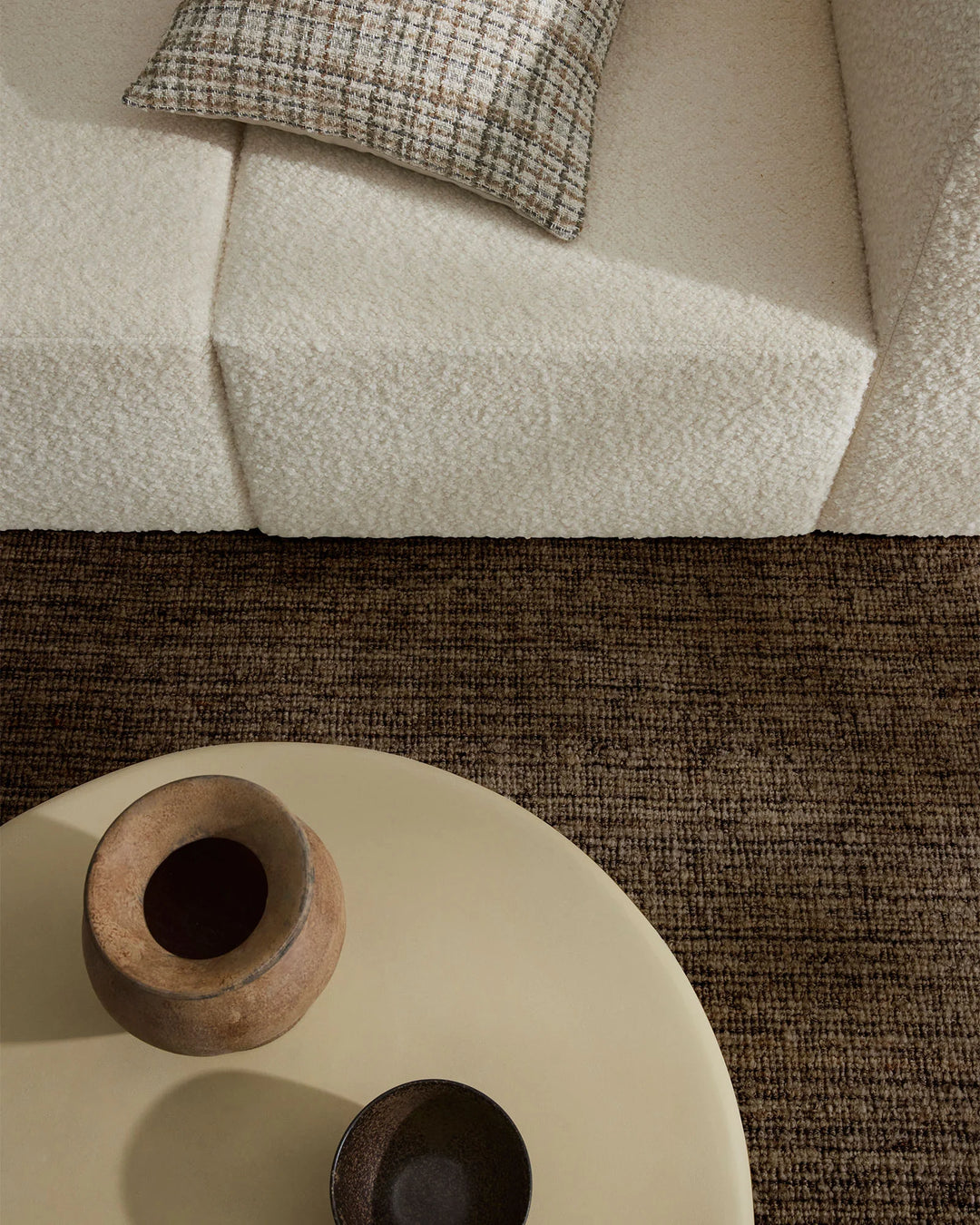 Neutral minimalist living room with cream and taupe textiles, featuring a textured Puglia rug in rich brown tones anchoring a white sofa with woven pillow and sculptural wood-base side table