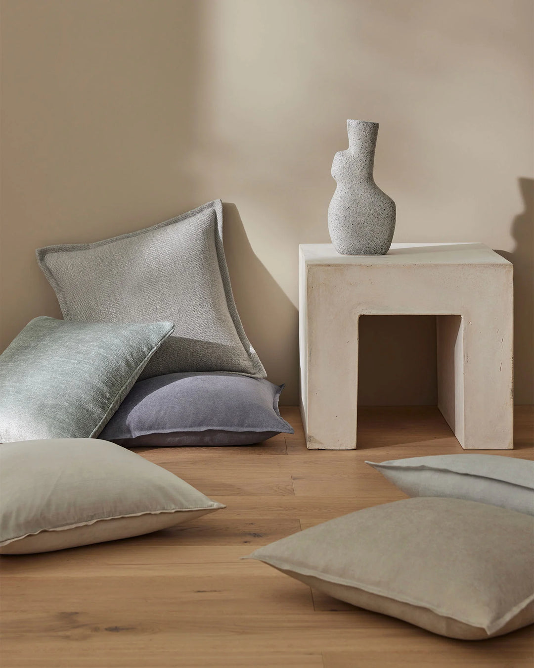 Neutral minimalist bedroom scene with layered linen cushions in sage, cream and slate blue tones arranged on wooden floor beside cream plaster side table
