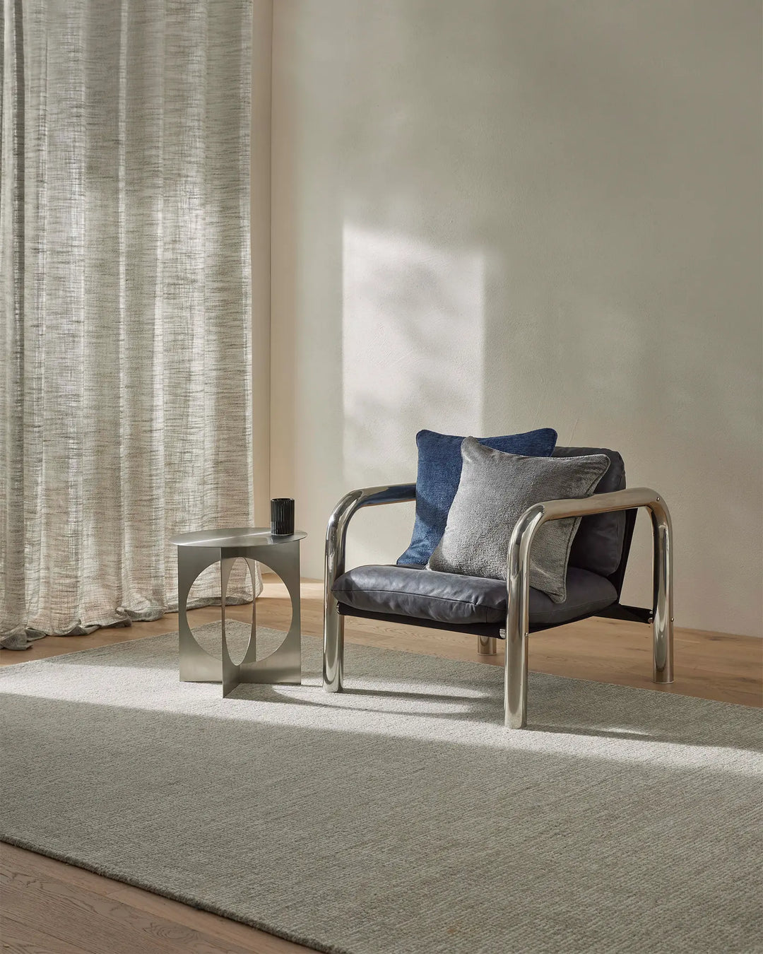 Minimalist living room with Kaia Mist rug in soft grey-beige, layered with linen curtains and navy cushions on a modern black leather chair, natural light streaming across neutral tones and wooden flooring