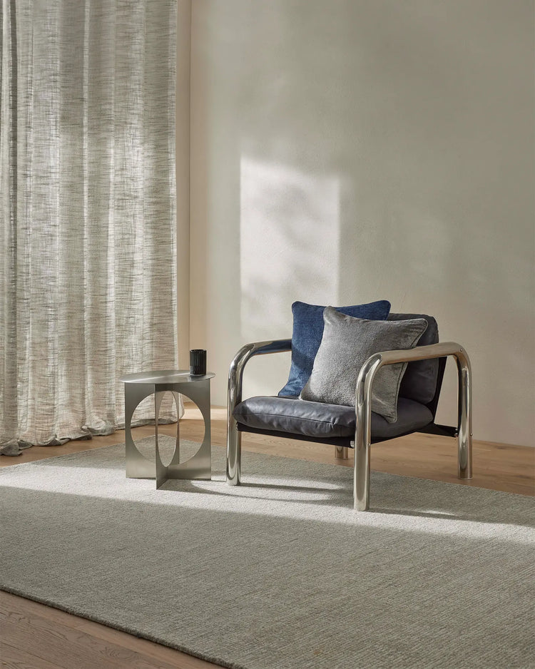 Minimalist living room with Kaia Mist rug in soft grey-beige, layered with linen curtains and navy cushions on a modern black leather chair, natural light streaming across neutral tones and wooden flooring