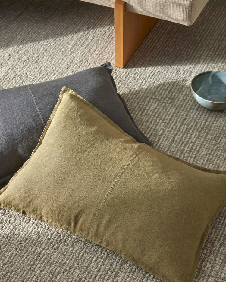 Warm sage linen cushion layered on charcoal upholstery with natural wool rug in neutral tones, styled in minimalist living room with timber furniture and soft daylight
