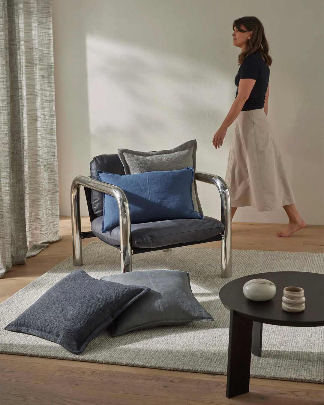 Minimalist living room styled with layered textiles: slate blue and charcoal cushions on a steel-framed chair, neutral wool rug, and natural linen curtain filtering soft light across warm timber flooring