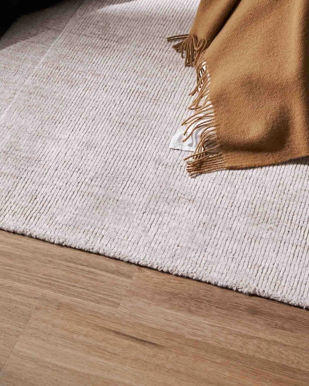 Camel wool throw draped over cream ribbed rug on light oak flooring, warm neutral earth tones with textured weave detail and fringed edges