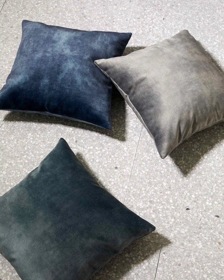 Three velvet cushions in navy, charcoal and soft grey arranged on light concrete, showcasing rich jewel-tone and neutral textures in contemporary minimalist styling