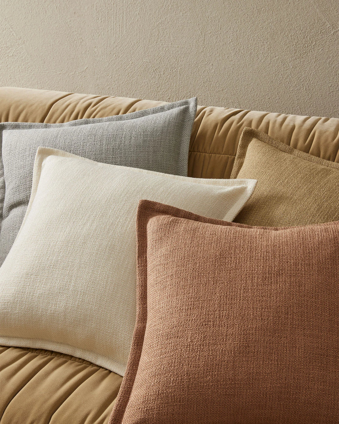 Neutral layered cushion styling on a quilted sofa, featuring cream, grey, terracotta and warm sand textured pillows against a textured beige wall