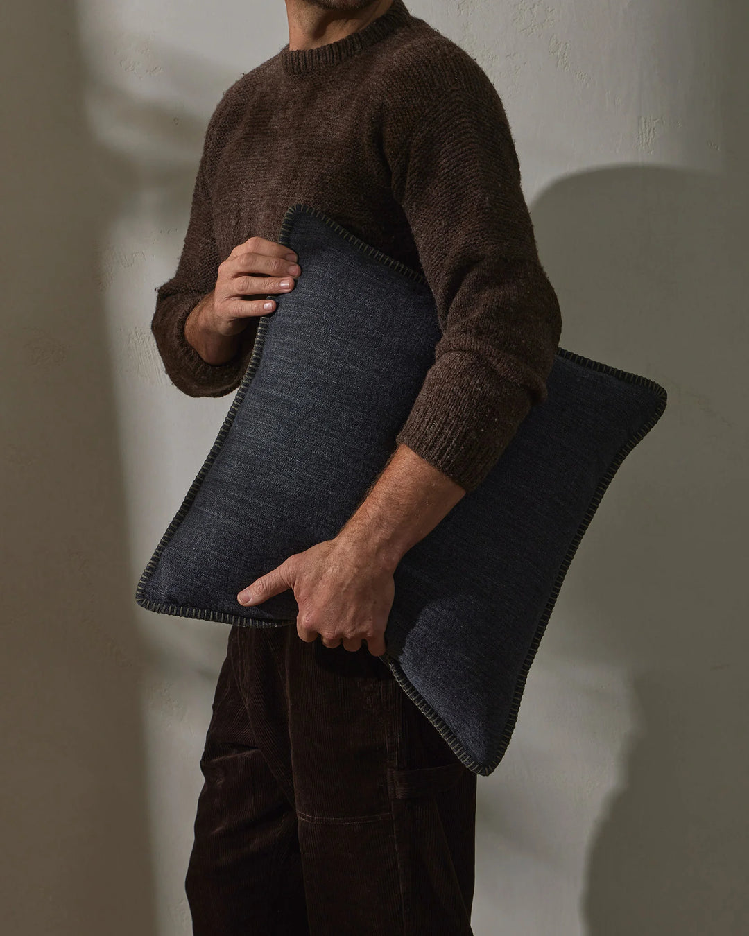 Man holding charcoal linen cushion against brown knit sweater, earthy colour palette with natural textures, minimalist styling against white backdrop