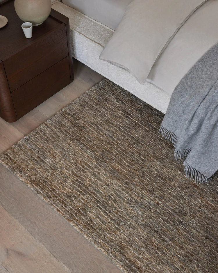 Neutral bedroom with Bastian Fossil jute rug anchoring white bedding and grey throw, dark wood nightstand, warm timber flooring, textured layering in stone and ash tones