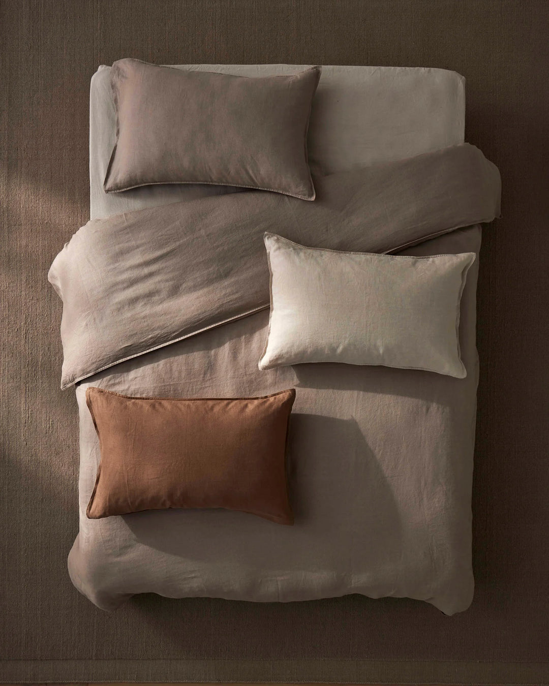 Overhead bed shot styled with warm terracotta and neutral linen bedding. Ravello flat sheet in pebble grey anchors the composition, layered with cream pillows and a Fiore cushion in rosewood for earthy tonal contrast.