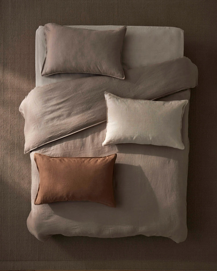 Overhead bed shot styled with warm terracotta and neutral linen bedding. Ravello flat sheet in pebble grey anchors the composition, layered with cream pillows and a Fiore cushion in rosewood for earthy tonal contrast.