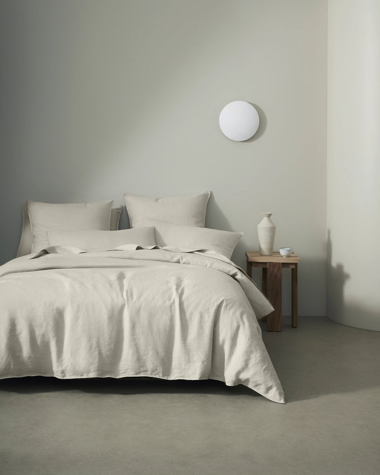 Minimalist bedroom styled with Ravello bedding in bone linen, featuring layered pillows and soft neutral textures against calm sage-green walls and wood nightstand