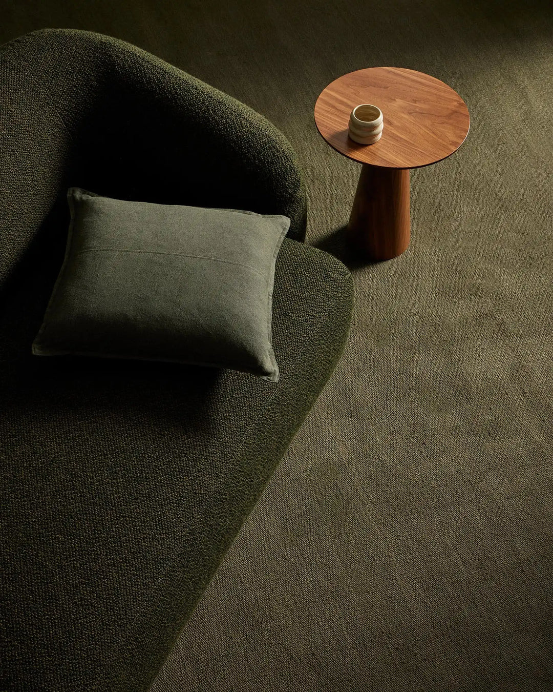 Sage green linen rug in modern living room with olive cushion on curved chair, warm wood side table, and natural light creating soft shadows