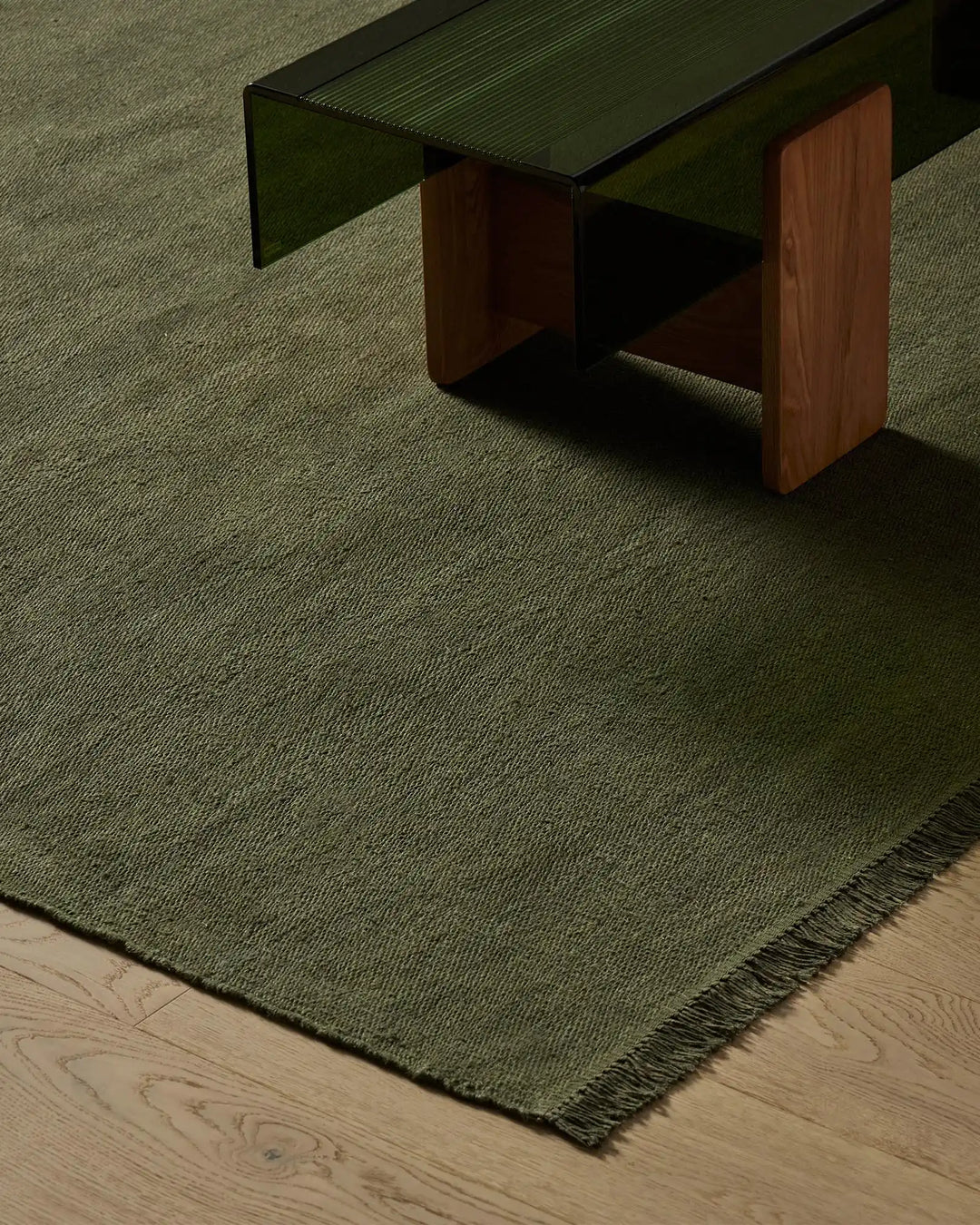 Sage green linen rug with subtle striped texture and fringed edge layered on light wooden flooring, styled under mid-century modern furniture in warm timber and forest tones