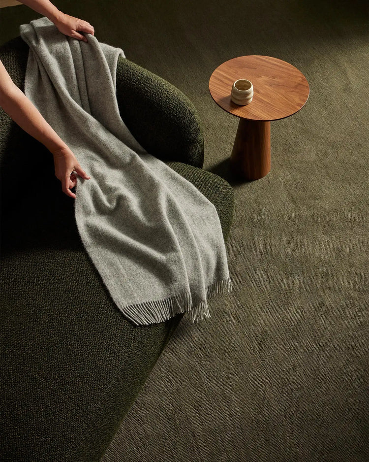 Neutral linen throw draped over green textured armchair with warm wood side table, earthy sage and ash palette, minimalist Scandinavian interior styling