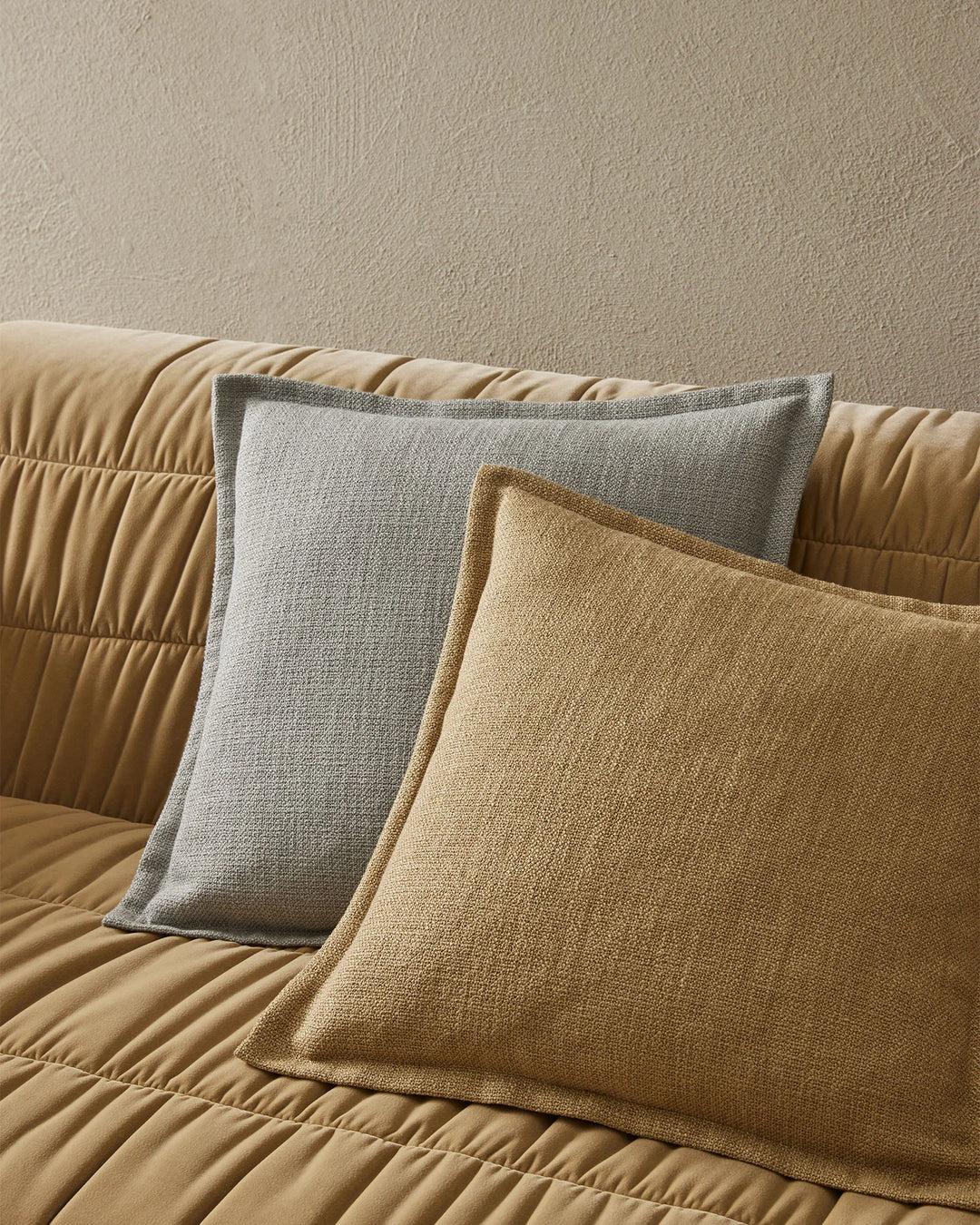 Neutral textured cushions in grey and warm gold tones arranged on a quilted caramel sofa against a soft taupe wall, showcasing layered minimalist home styling