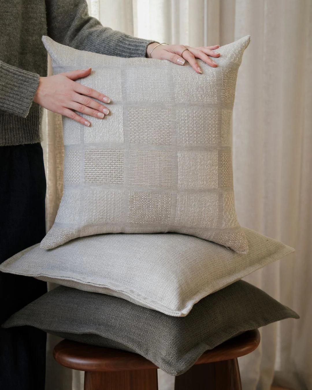 Neutral texture stack featuring cream patchwork and soft grey cushions on timber stool, styled with charcoal knit and warm natural light