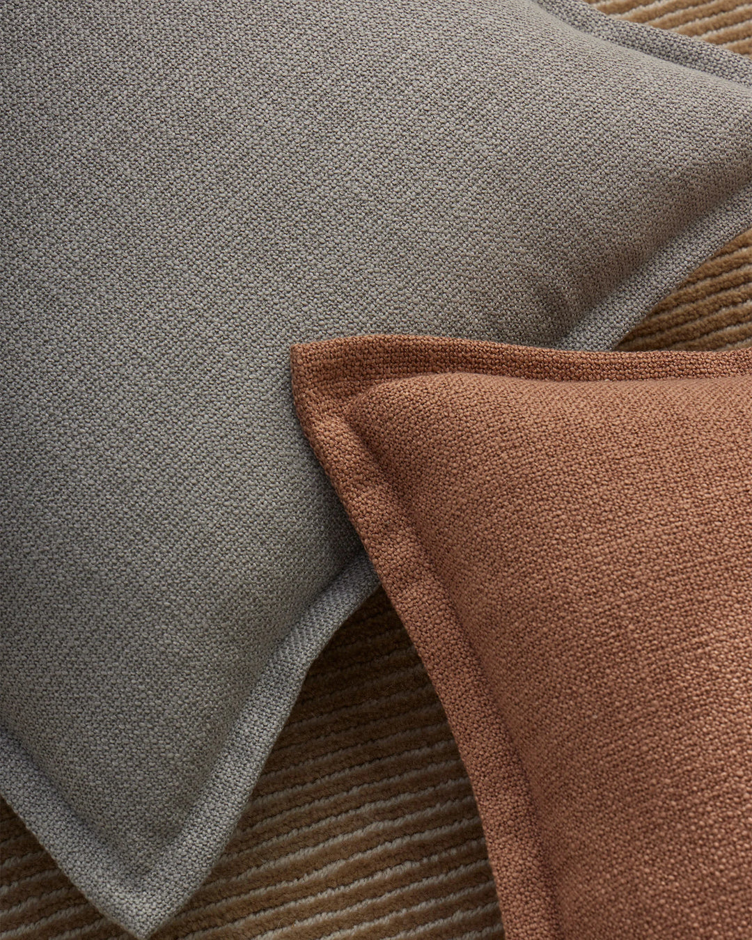 Layered Figlio cushions in mineral grey and terra tones display textured woven fabrics in warm, earthy neutrals with subtle depth and dimension
