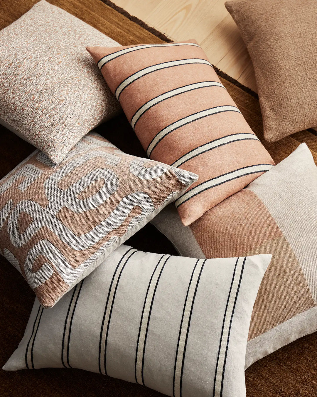 Warm terracotta and cream striped cushions styled with textured neutral pillows on wooden floorboards; earthy minimalist décor with linen and cotton blend fabrics