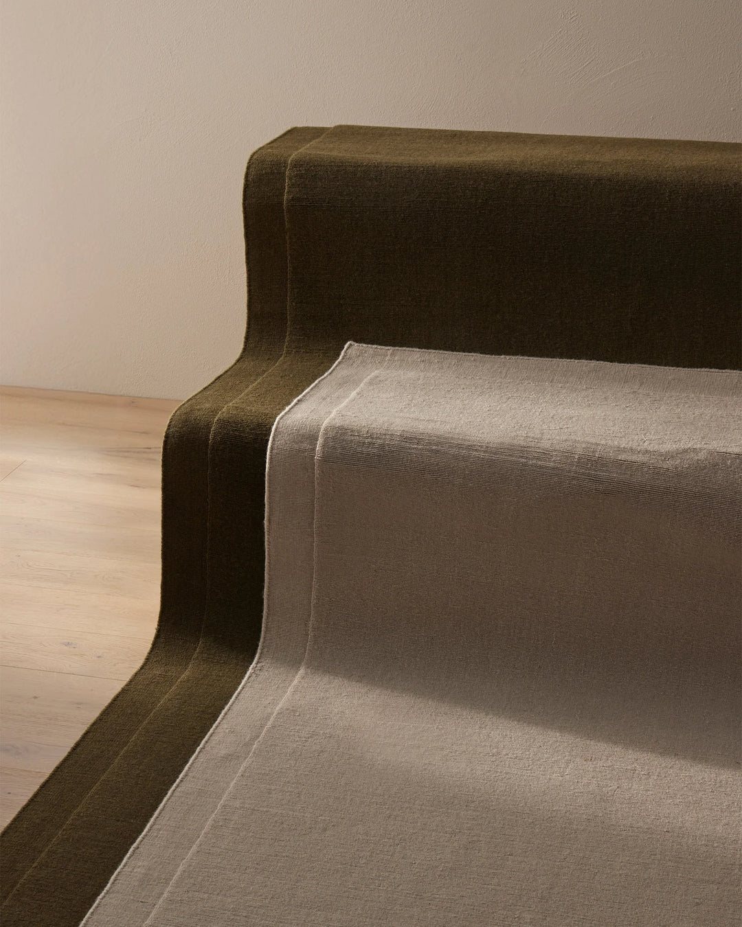 Earthy wool rugs in olive and soft grey layered on natural timber flooring, showcasing warm neutral tones and natural texture against minimalist walls