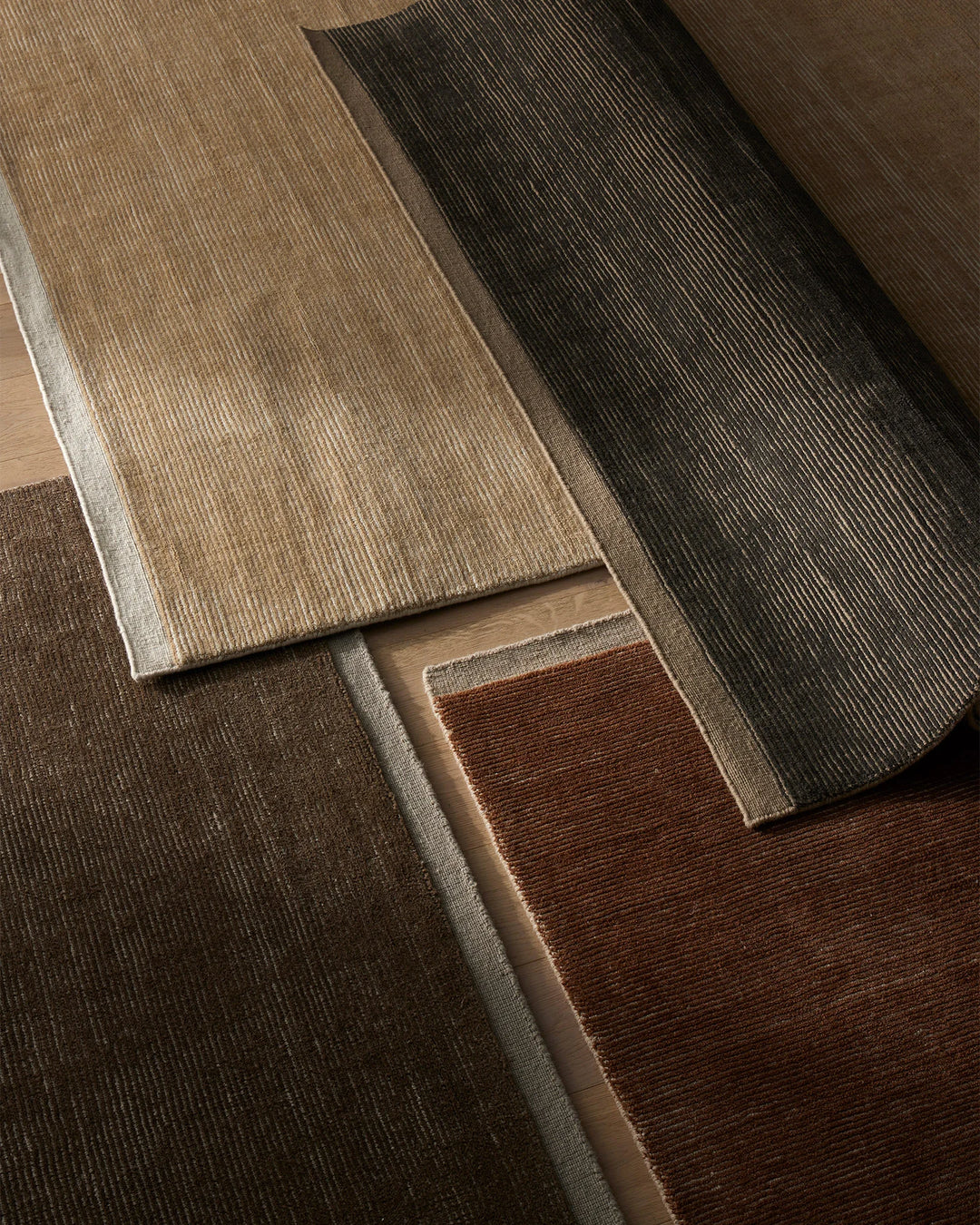 Overhead view of neutral and earthy wool rugs layered together, showcasing textured ribbed weaves in sand, charcoal, terracotta and warm brown tones with soft natural light