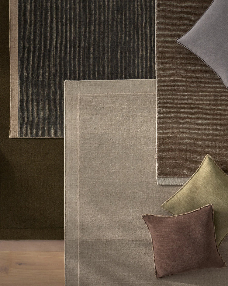 Layered wool rugs in earth tones and charcoal create a textured floor styling; neutral palette with sage, taupe, and warm brown tones featuring cushions with whipstitched detailing