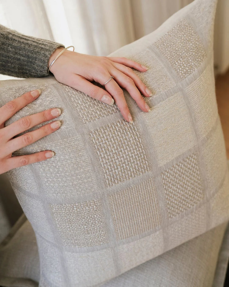 Neutral linen cushion with geometric weave texture held against cream and charcoal layered textiles, showcasing tactile woven patterns and earthy minimalist styling