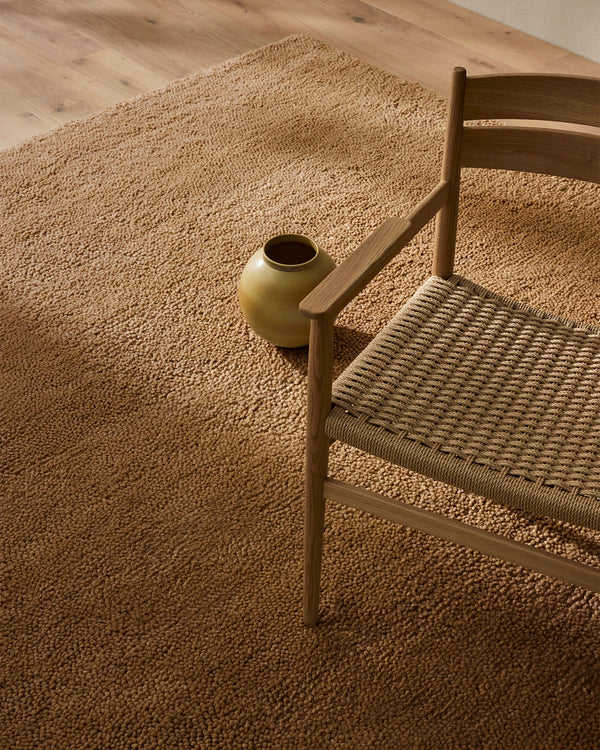 Neutral minimalist bedroom with Valencia Camel wool rug in warm sand tone, wooden chair with woven seat, ochre pottery vessel, natural light casting soft shadows on textured flooring