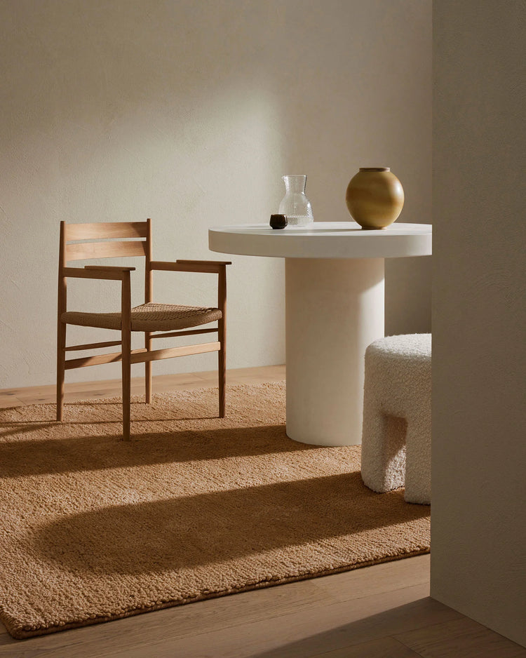 Warm minimalist interior with camel wool rug layered under sculptural furniture, natural beige and cream palette with woven textures and soft afternoon light