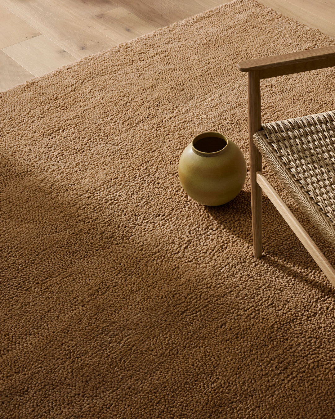 Warm neutral living room with textured camel wool rug, wooden chair with patterned upholstery, and ochre ceramic vessel on light timber flooring