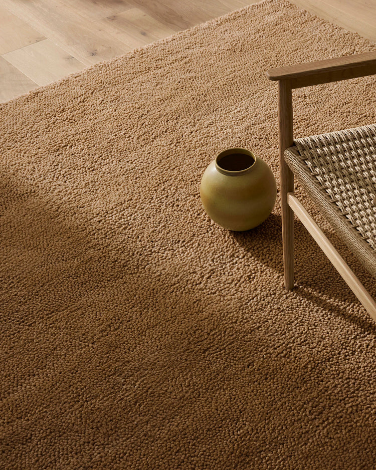 Warm neutral living room with textured camel wool rug, wooden chair with patterned upholstery, and ochre ceramic vessel on light timber flooring
