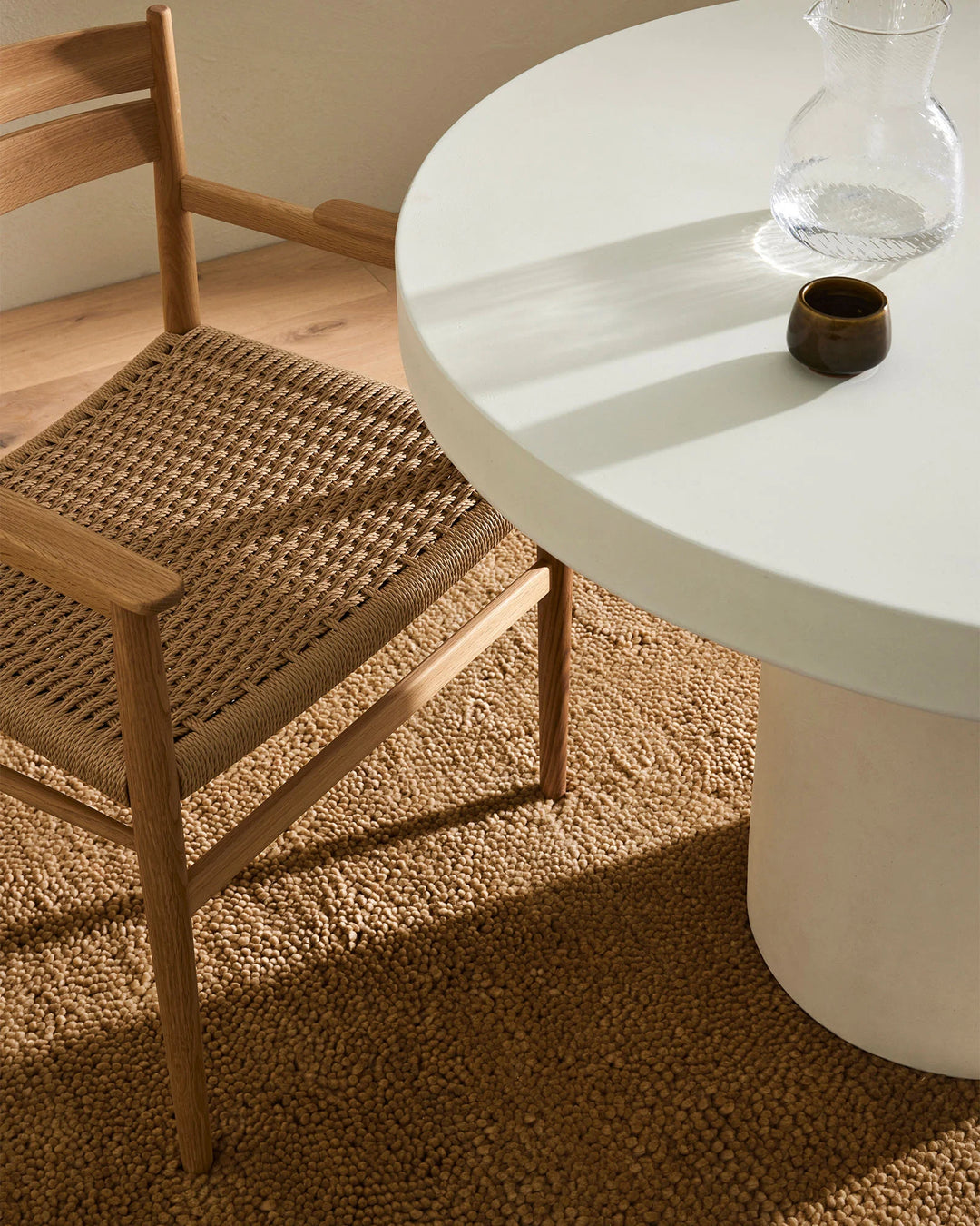 Warm neutral dining scene with woven wood chair, textured camel wool rug, white round table, and natural light casting soft shadows