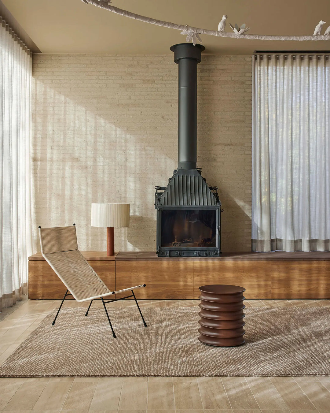 Neutral mid-century living room with natural linen curtains, warm timber tones, and a textured wool rug anchoring the space beside a black stove