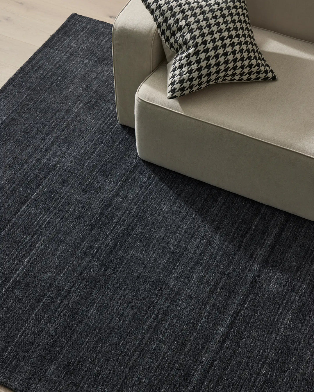 Charcoal textured rug anchors a minimalist seating vignette with cream sofa and houndstooth cushion, showcasing neutral monochromatic styling