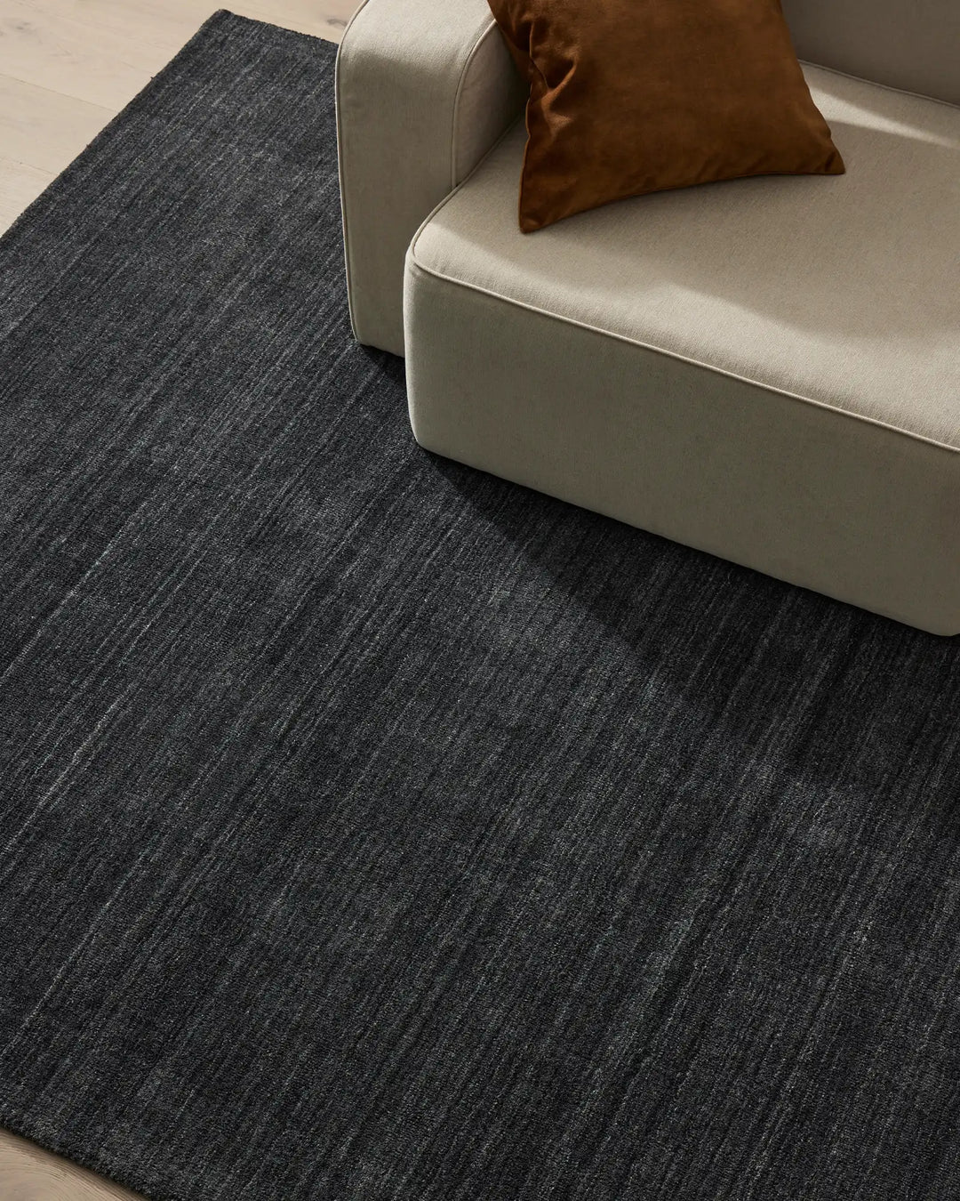 Charcoal textured Gippsland rug anchors minimalist living room with cream sofa and cognac leather cushion, cool neutral palette with warm accents