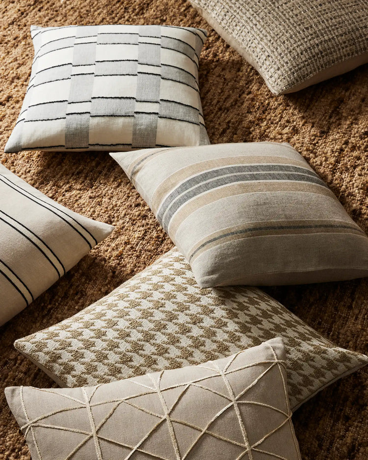 Neutral linen and cotton cushions in cream, charcoal and taupe tones layered on a textured caramel rug, featuring geometric stripes, quilted patterns and woven details