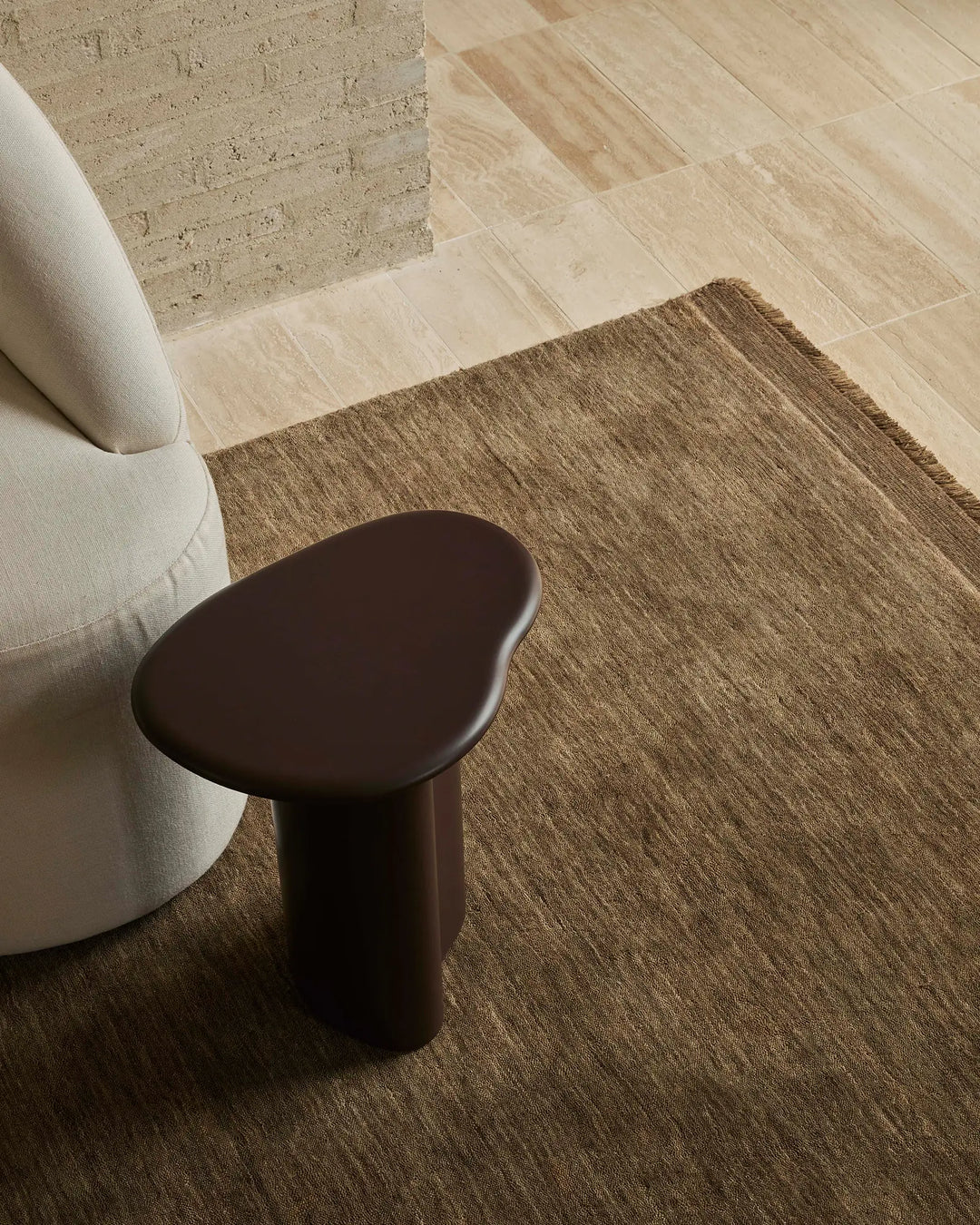 Neutral minimalist interior with warm taupe area rug, cream curved chair, and dark brown side table on light wood flooring beside textured brick wall