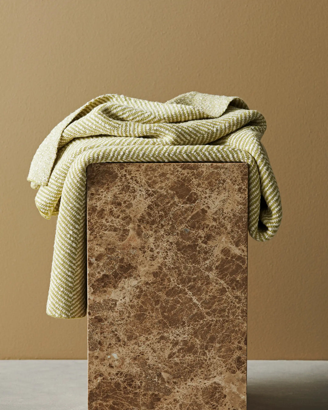 Olive and cream striped cotton throw casually draped over a speckled brown marble plinth, warm neutral palette against soft tan backdrop
