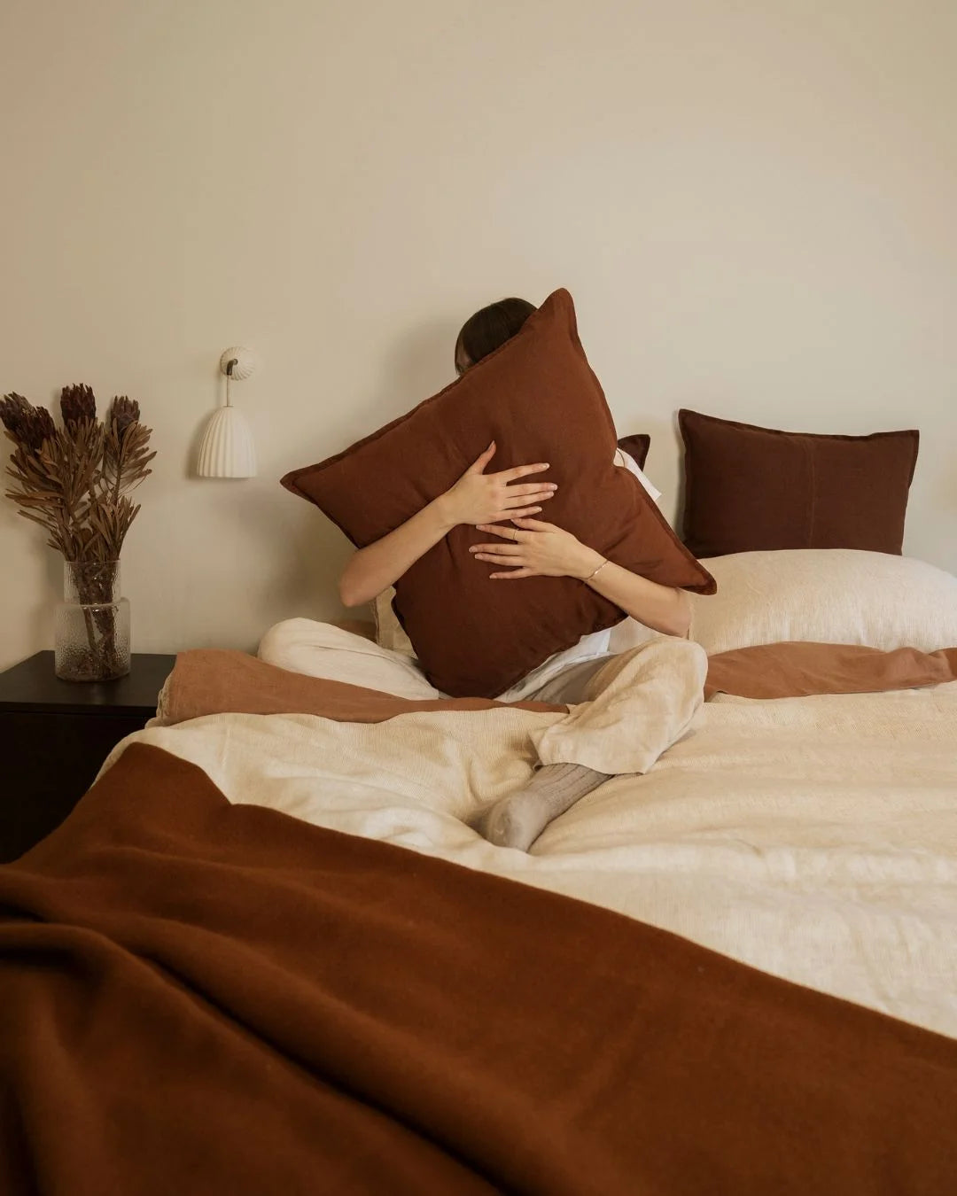 Warm earth-tone bedroom with chocolate brown linen cushions, spiced wool throw, and cream pinstriped bedding layered for texture and comfort
