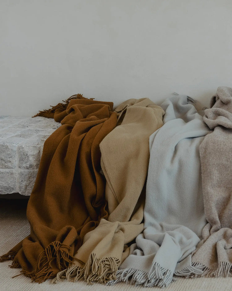 Nevis Spice lambswool throw in burnt orange-brown with tassel fringe, displayed among neutral wool throws by Weave Home