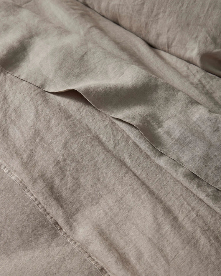 Neutral linen bedding in warm pebble tones with soft texture and gentle folds, showcasing the Ravello Flat Sheet's lightweight European linen weave and relaxed, undone aesthetic