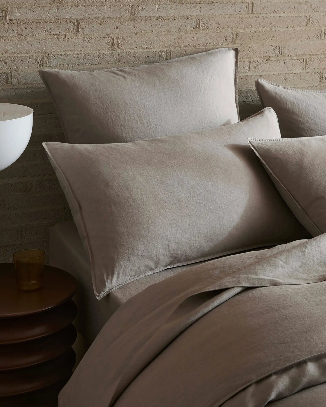 Neutral linen bedding styled on a bed with textured brick wall, featuring layered Ravello flat sheet in warm pebble tones and soft furnishings for minimalist bedroom aesthetic