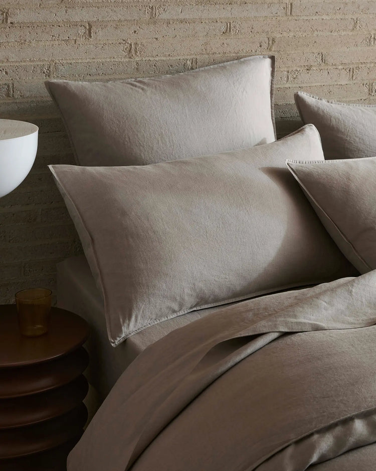 Neutral linen bedding styled on a bed with textured brick wall, featuring layered Ravello flat sheet in warm pebble tones and soft furnishings for minimalist bedroom aesthetic