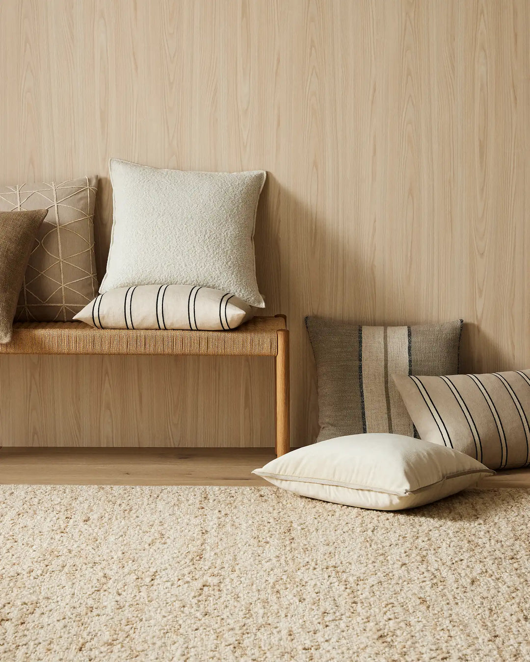 Neutral linen and linen-blend cushions in cream, taupe and striped patterns arranged on timber bench with natural jute rug, warm wood paneling backdrop