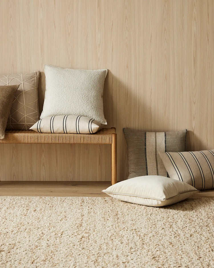 Neutral linen and linen-blend cushions in cream, taupe and striped patterns arranged on timber bench with natural jute rug, warm wood paneling backdrop
