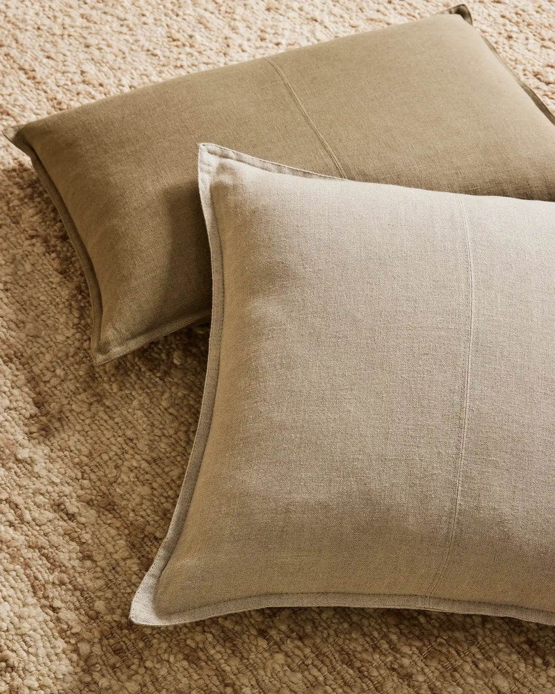 Neutral linen cushions layered on textured wool rug, soft earth-tone palette with warm beige and sage tones, minimal modern styling with natural fabric texture